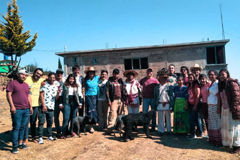 Large group of people standing outdoors in front of a building with two black dogs in the foreground.