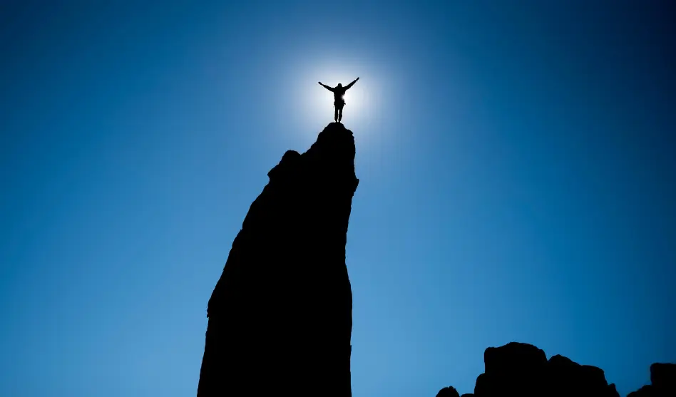 Silhouette of a person standing with arms raised on top of a tall rock formation, backlit by the sun against a clear blue sky.