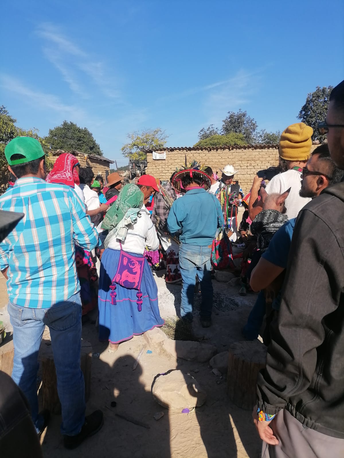 Group of people gathered in a village setting under a clear blue sky, wearing traditional clothing and feathered hats during a cultural ceremony.
