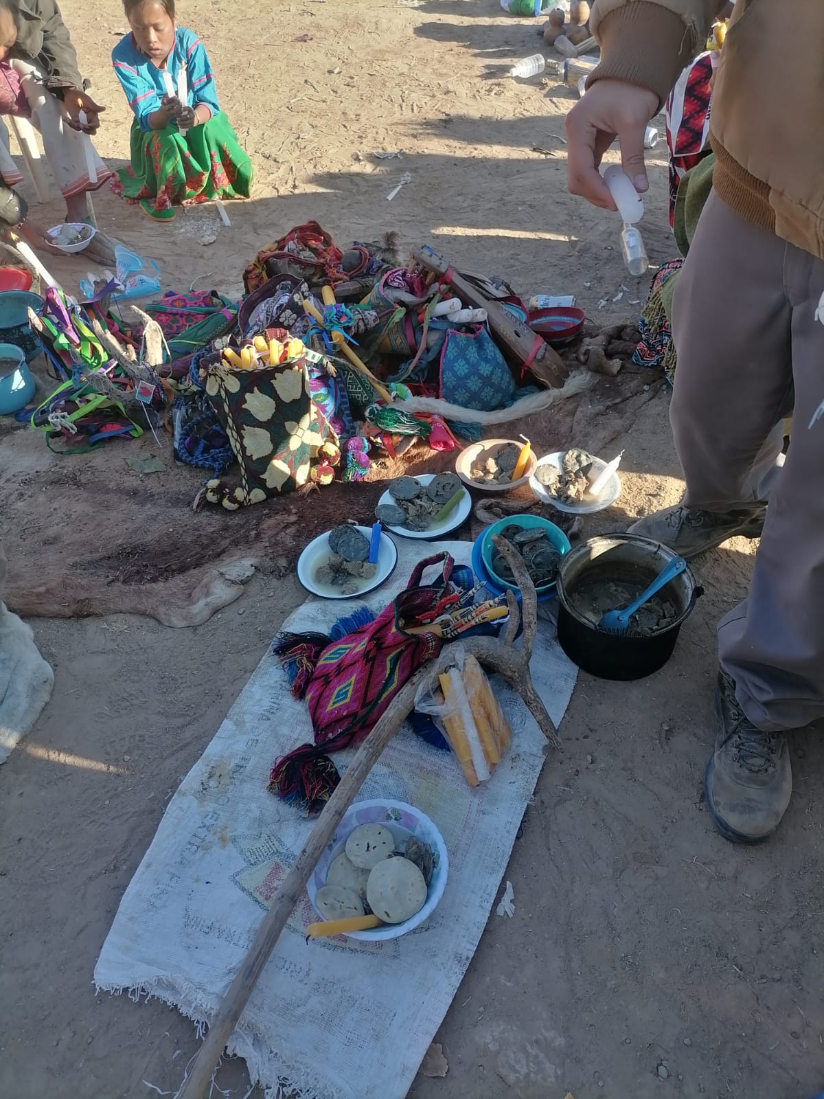 Outdoor cultural setup with traditional textiles, bowls of food, candles, and ritual objects arranged on a cloth mat on sandy ground, with people gathered nearby.