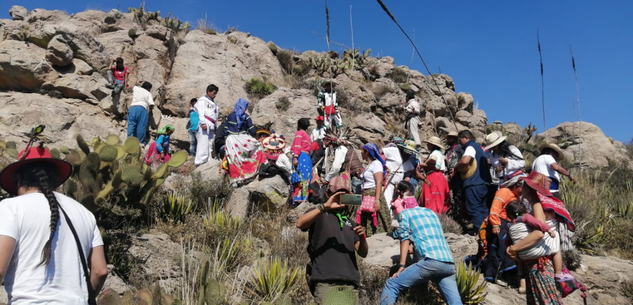 Group of people in colorful traditional clothing gathered on a rocky hillside with cacti under a clear blue sky, participating in an outdoor cultural event.