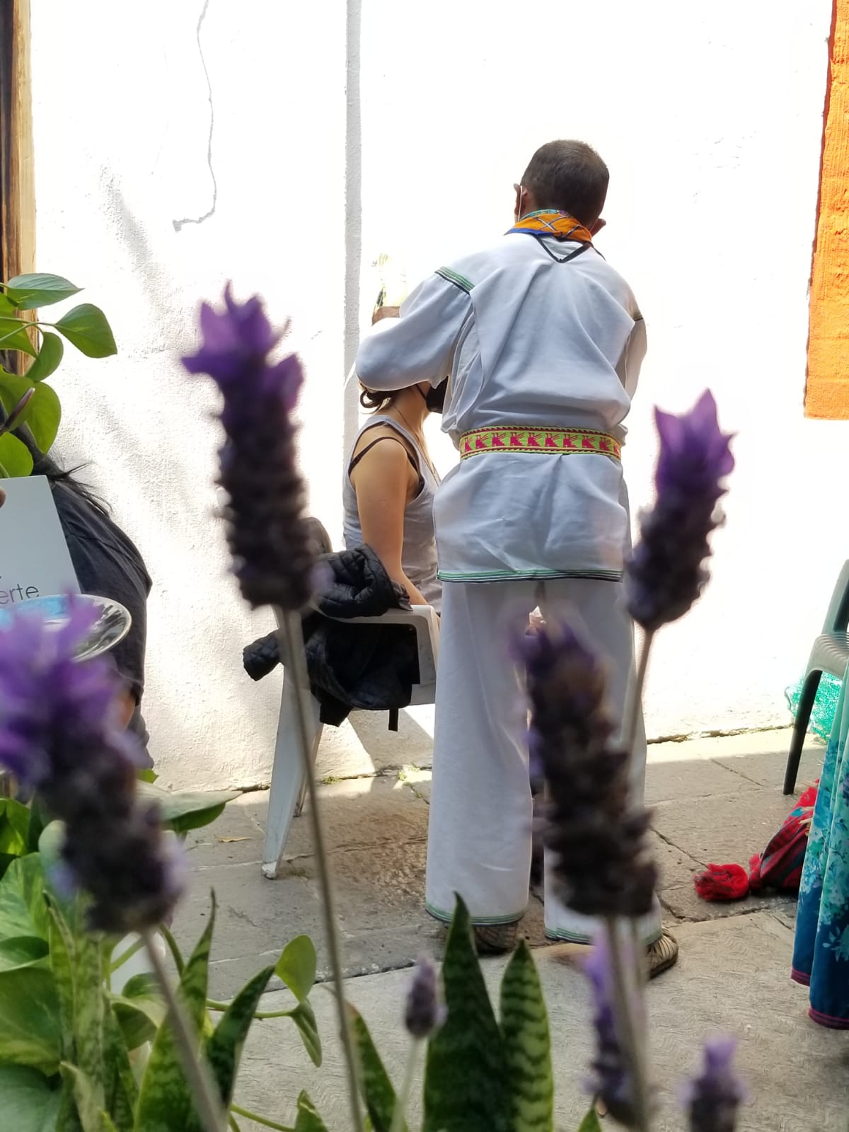Person in traditional attire standing beside a seated individual in an outdoor courtyard, with purple flowers blurred in the foreground.