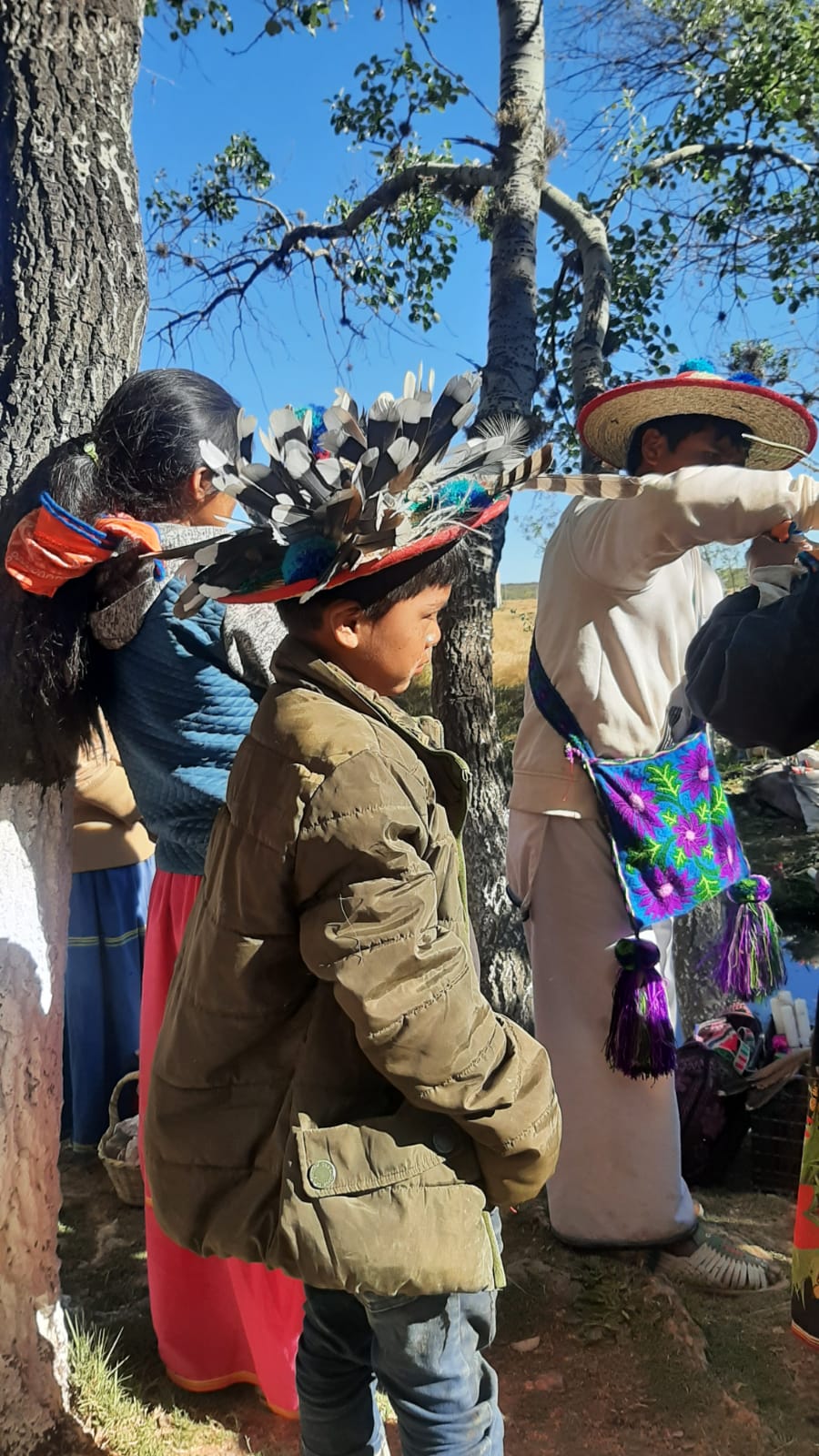 Child and adults wearing traditional clothing and feathered hats standing outdoors near trees during a cultural gathering.