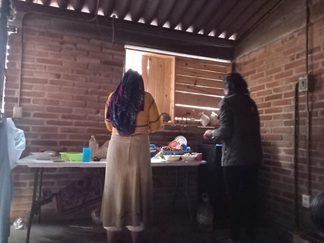 Two women preparing food inside a rustic brick kitchen, standing at a table with dishes and utensils near a wooden window.
