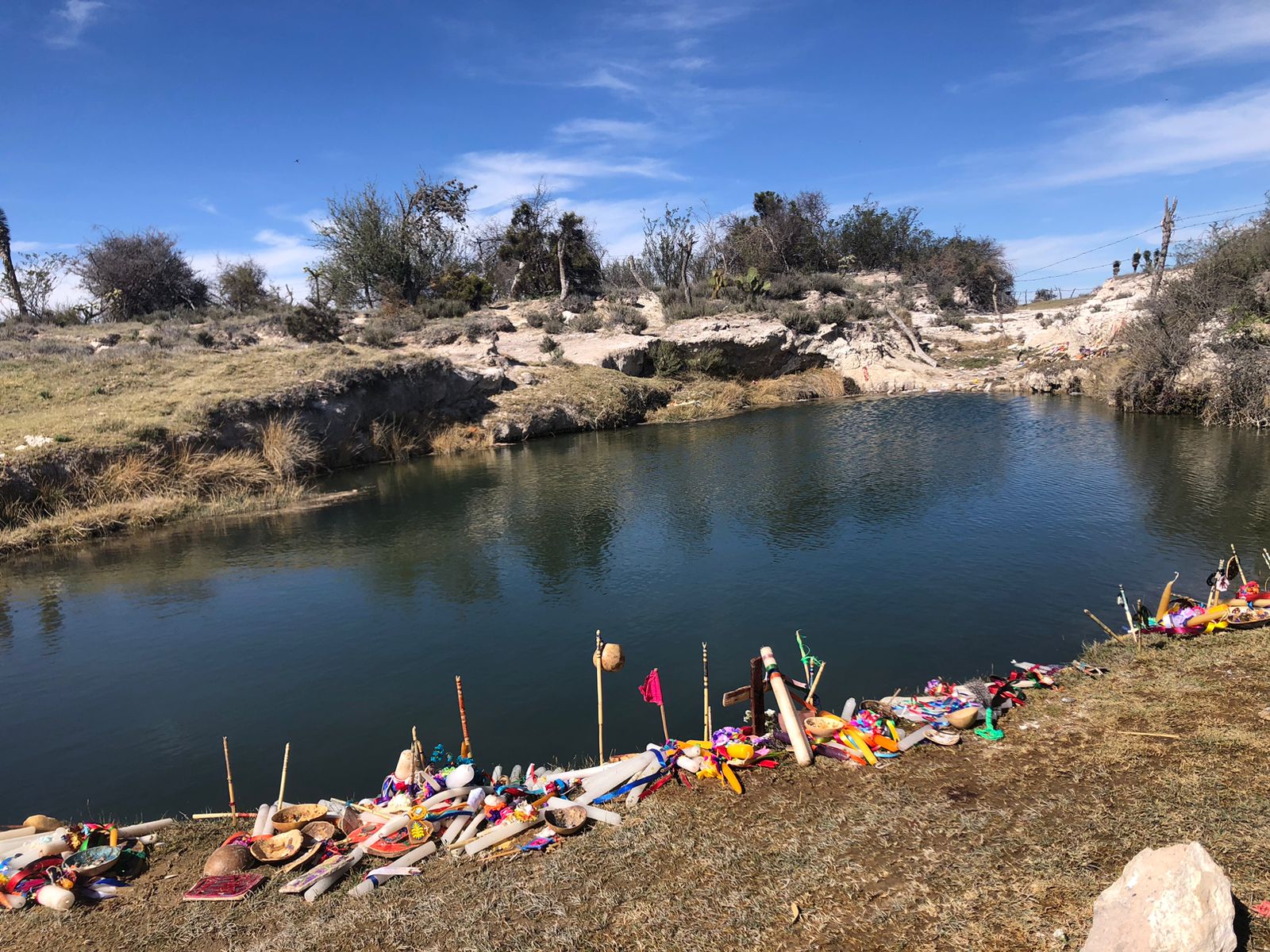 Calm river surrounded by rocky terrain and sparse trees, with colorful ceremonial objects arranged along the riverbank under a clear blue sky.