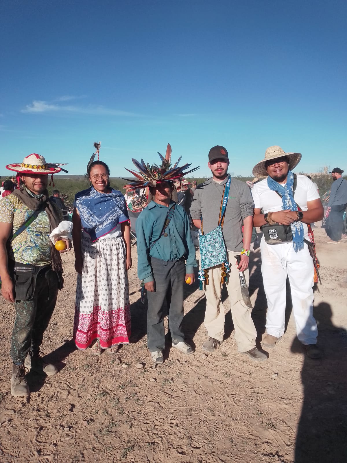 Five people standing outdoors in a desert landscape wearing traditional clothing and feathered headdresses, posing together during a cultural gathering under a clear blue sky.