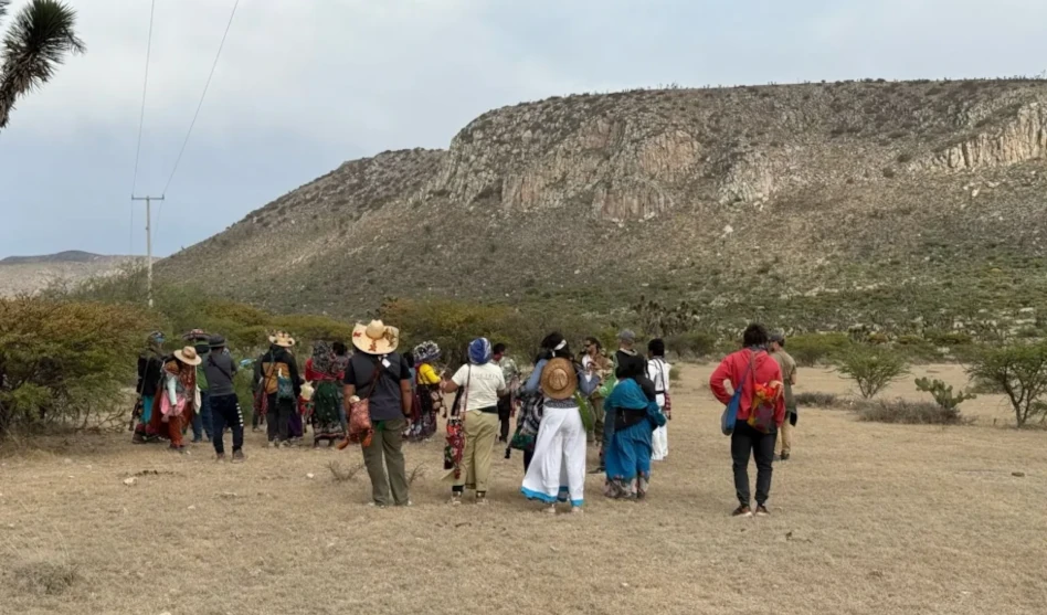 Group of people walking across a dry open landscape toward a rocky hill, wearing traditional clothing and hats during an outdoor cultural gathering.