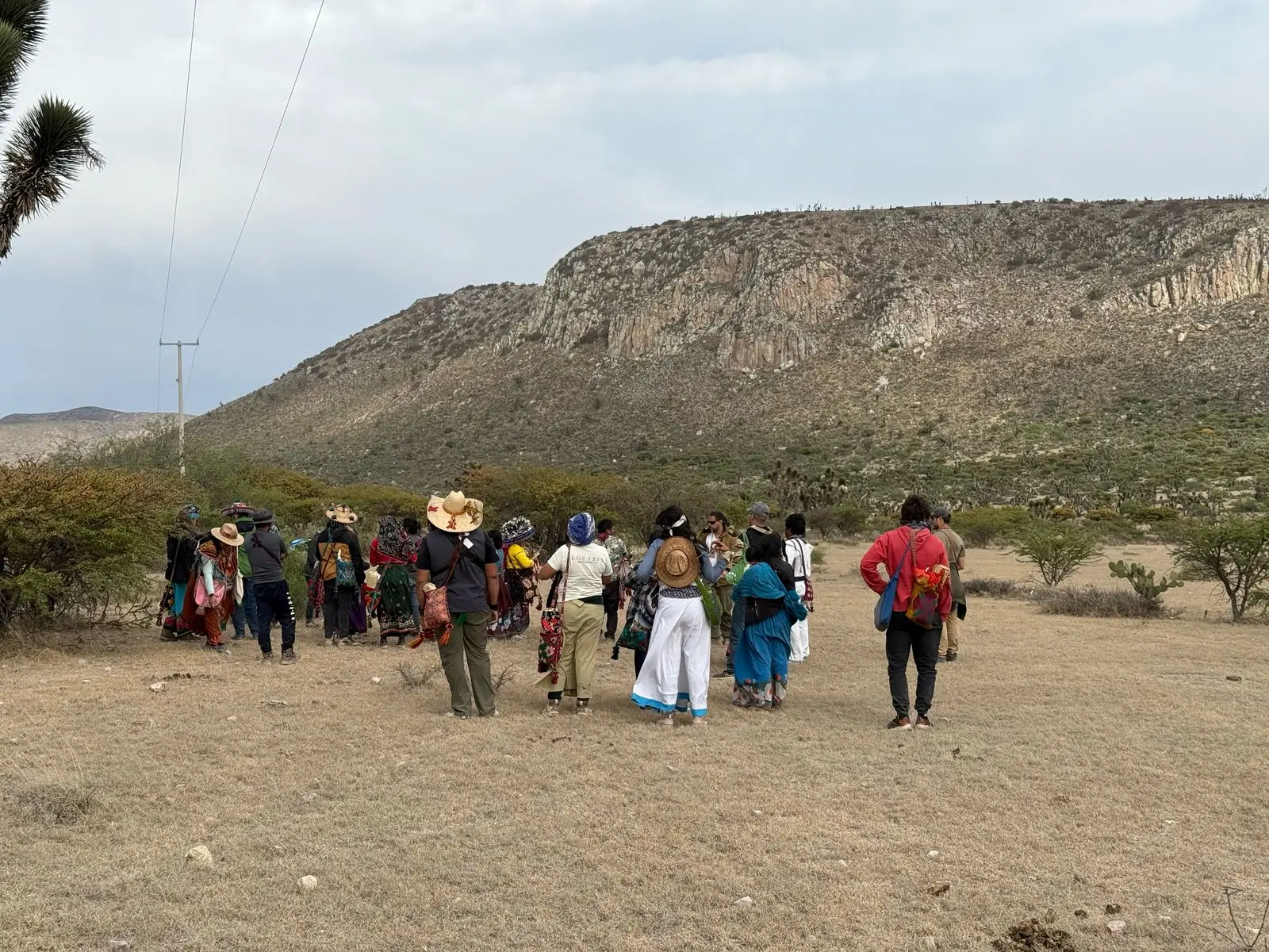 Group of people walking across a dry open field toward a rocky hillside, wearing traditional clothing and hats during an outdoor cultural gathering.