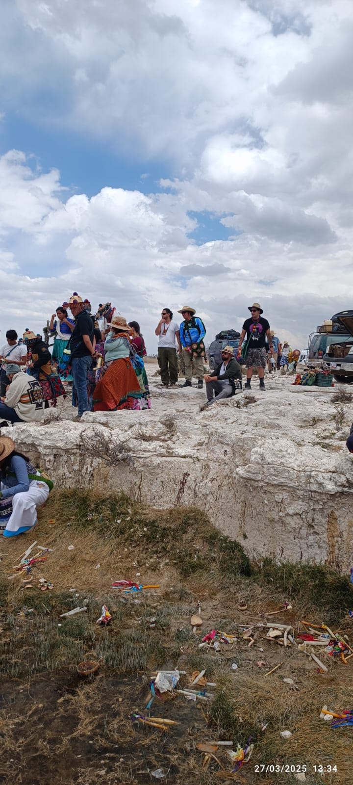 Group of people gathered on a rocky plateau under a cloudy sky during an outdoor cultural event, with traditional clothing, vehicles, and ceremonial items visible nearby.