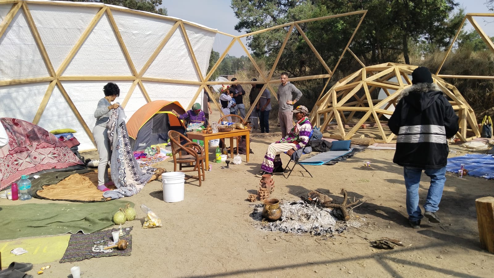 People gathered at an outdoor campsite with geometric dome structures, tents, and a small fire pit, participating in a cultural or community event.