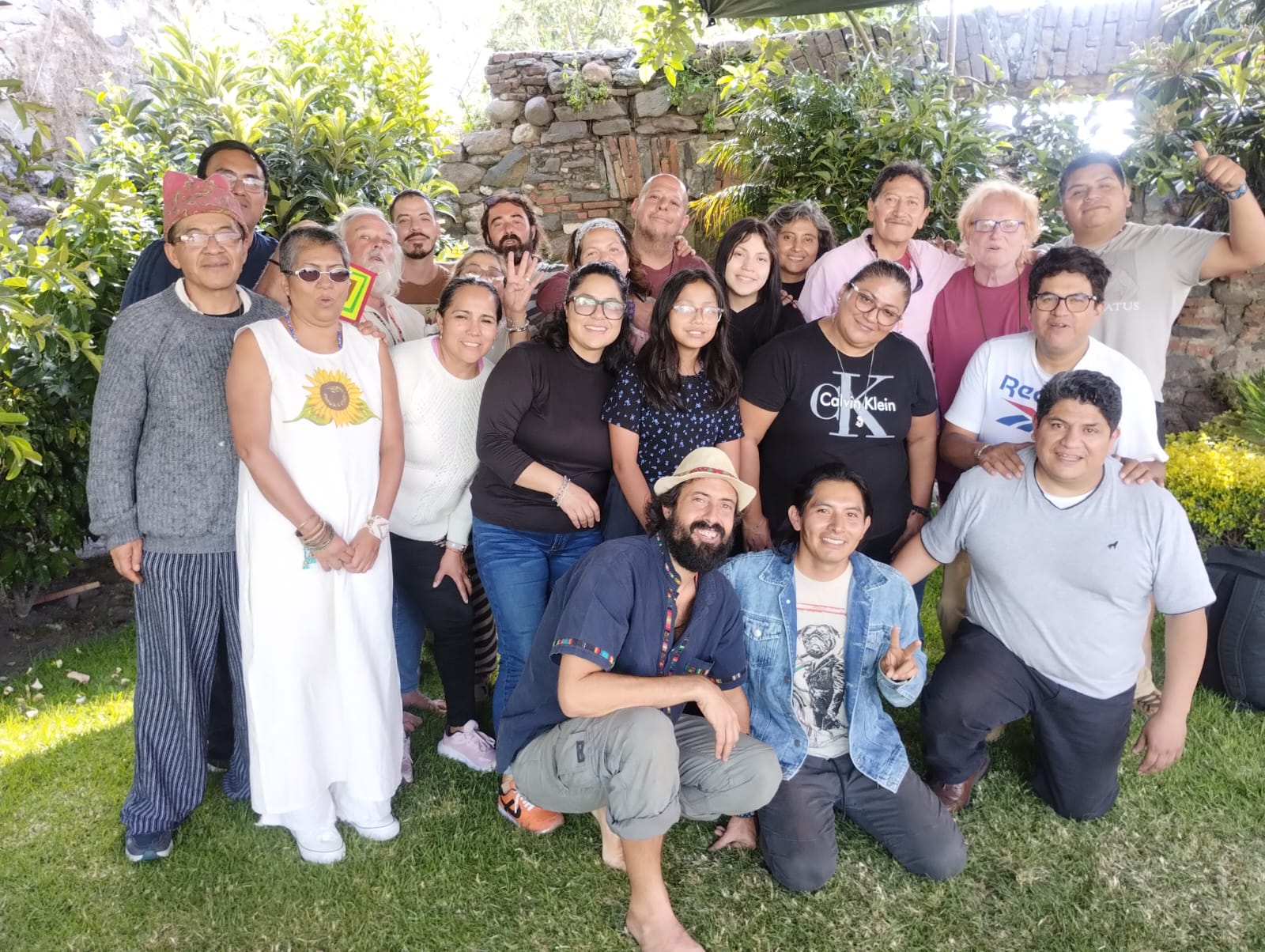 Large group of people smiling and posing together outdoors in a garden setting with greenery and a stone wall in the background.