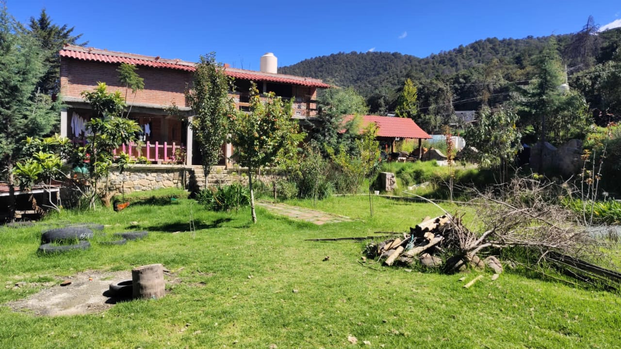 Rustic countryside home with a red tile roof surrounded by green lawn, trees, and forested hills under a clear blue sky.