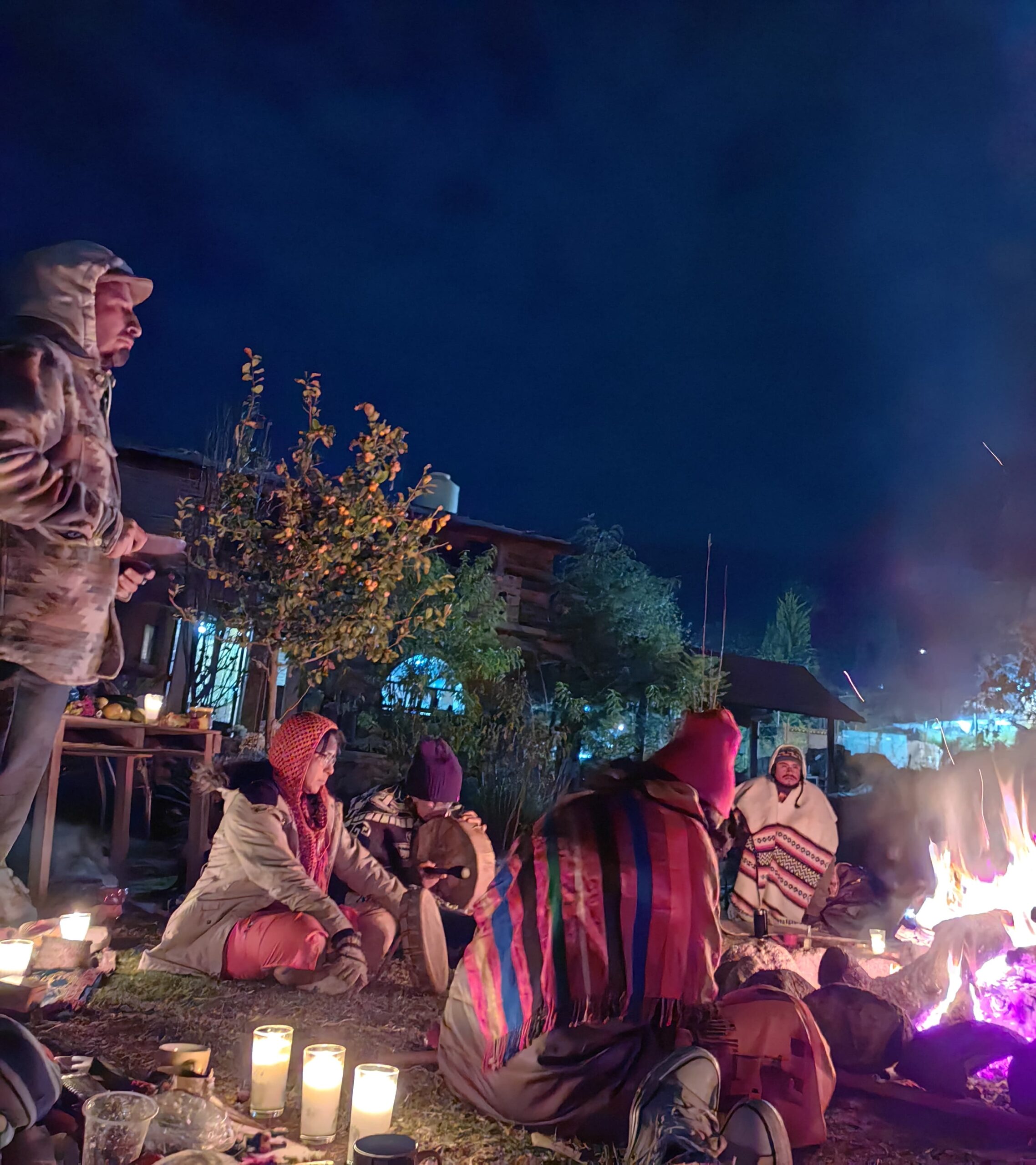 People gathered around a nighttime bonfire outdoors, wrapped in blankets and traditional clothing, with candles placed on the ground and a house visible in the background.