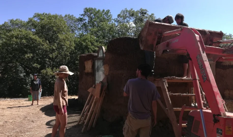 Several people working together on an earthen building structure outdoors, using a small lift and ladder under a clear blue sky with trees in the background.