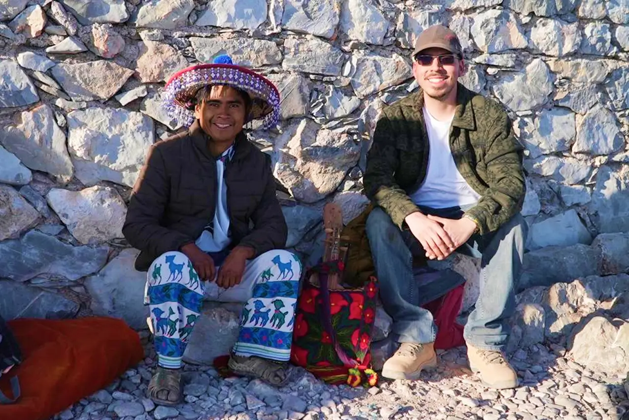 Two men sitting against a stone wall outdoors, smiling at the camera, one wearing traditional clothing and a colorful hat, the other in casual modern attire.