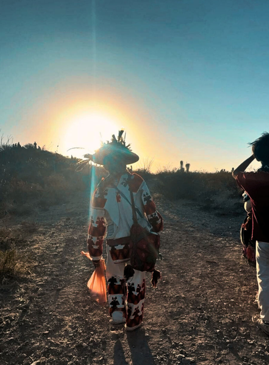Person in ceremonial attire holding a staff or torch at sunset in an open field.