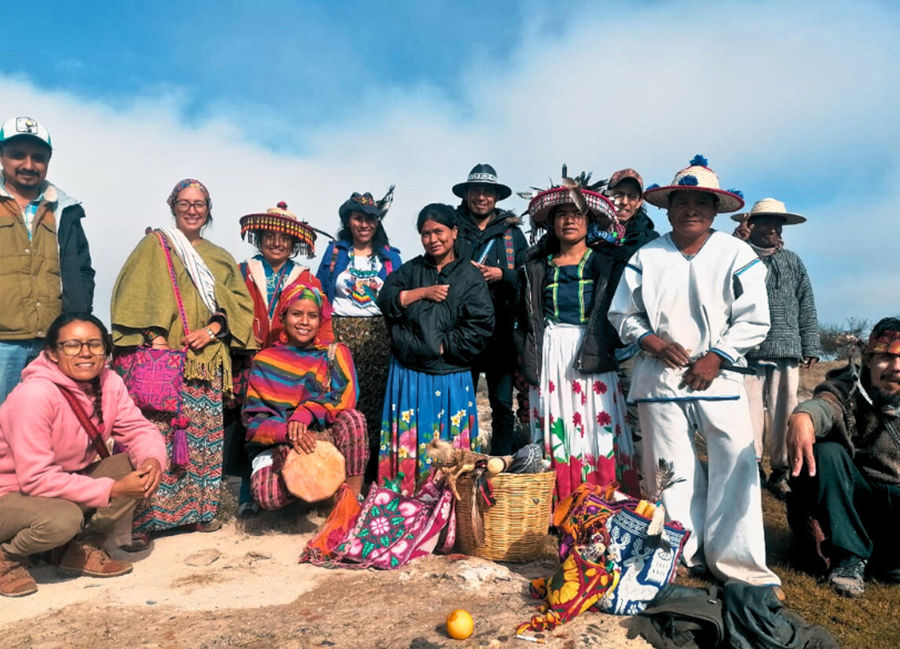 Group of men and women in traditional ceremonial clothing standing together outdoors with ritual offerings placed on the ground.